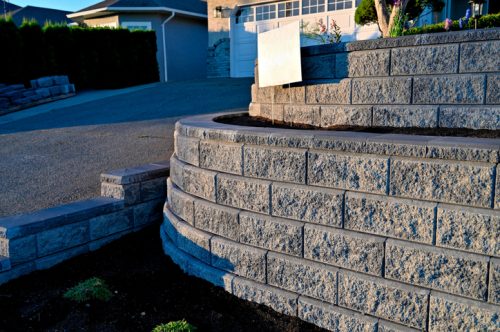 A close-up view from the side looking at the return of a two tier retaining wall returning in a curve back into the hill in gray color back lit by early morning sunlight.